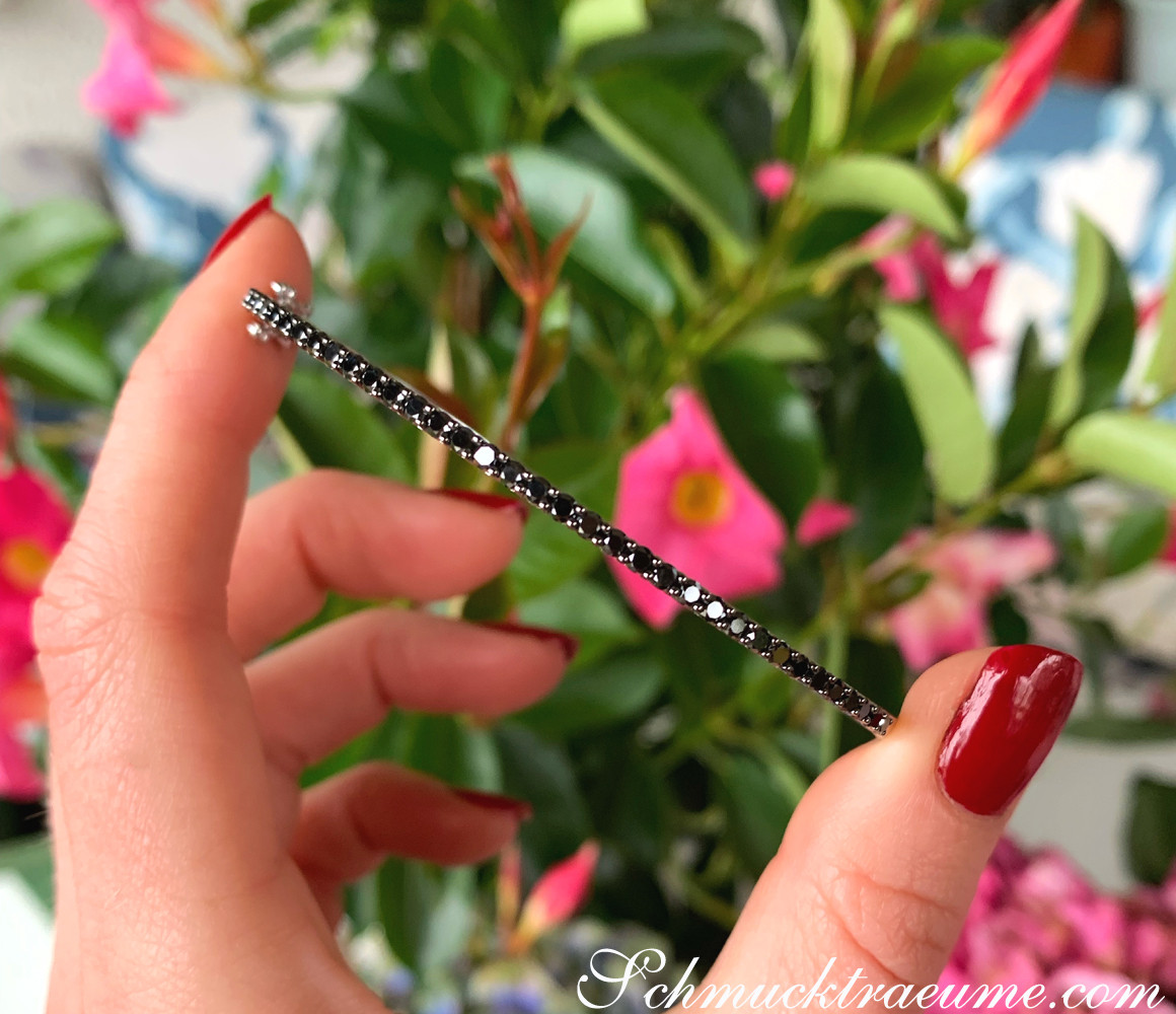 Weißgold-Armreif mit schwarzen Diamanten, Detailaufnahme, rote Nagellack, Blüten im Hintergrund.