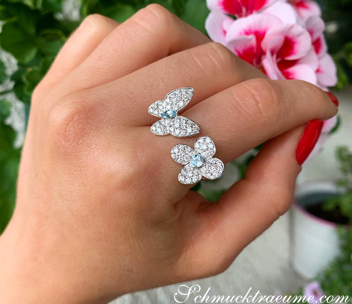 Finger mit brillantengeschmücktem Schmetterling-Ring vor Blütenhintergrund