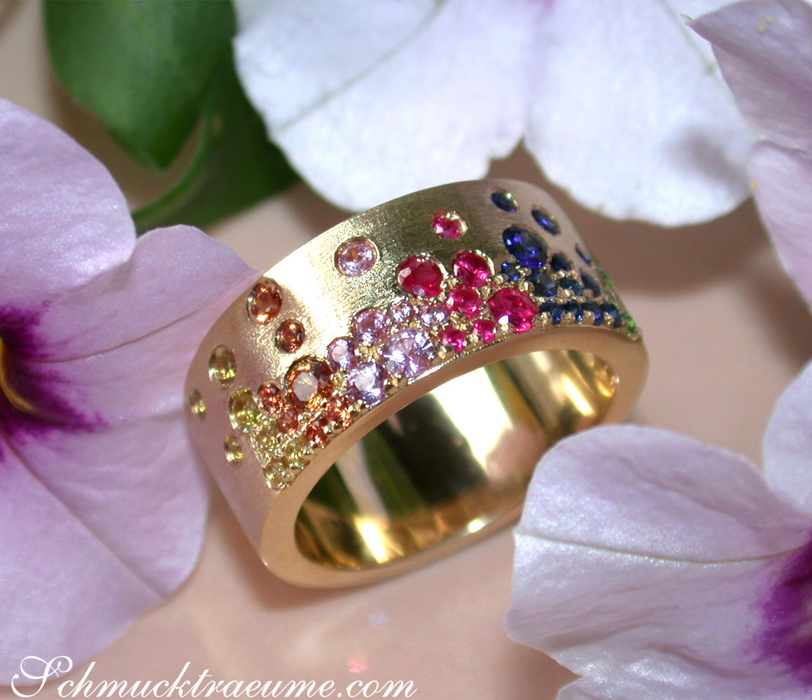 Goldener Ring mit farbigen Saphiren, Rubinen und Tsavorites, Detailaufnahme, satiniertes Gelbgold, Blüten im Hintergrund