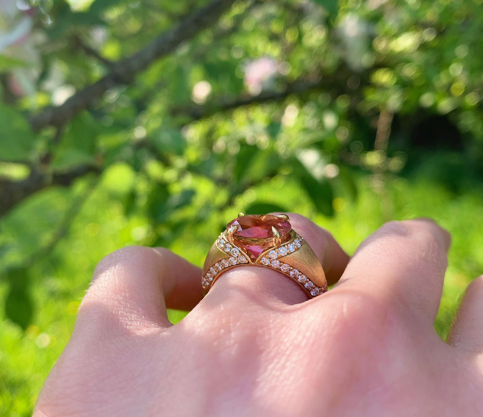 Nahaufnahme von Ring mit rosa Turmalin und Diamanten, im Hintergrund grüne Natur.