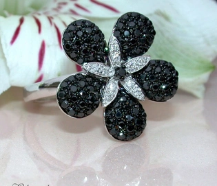 Blumenring mit schwarz-diamantenbesetzten Blütenblättern auf weißgoldigem Ring, vor Blütenblättern im Hintergrund.