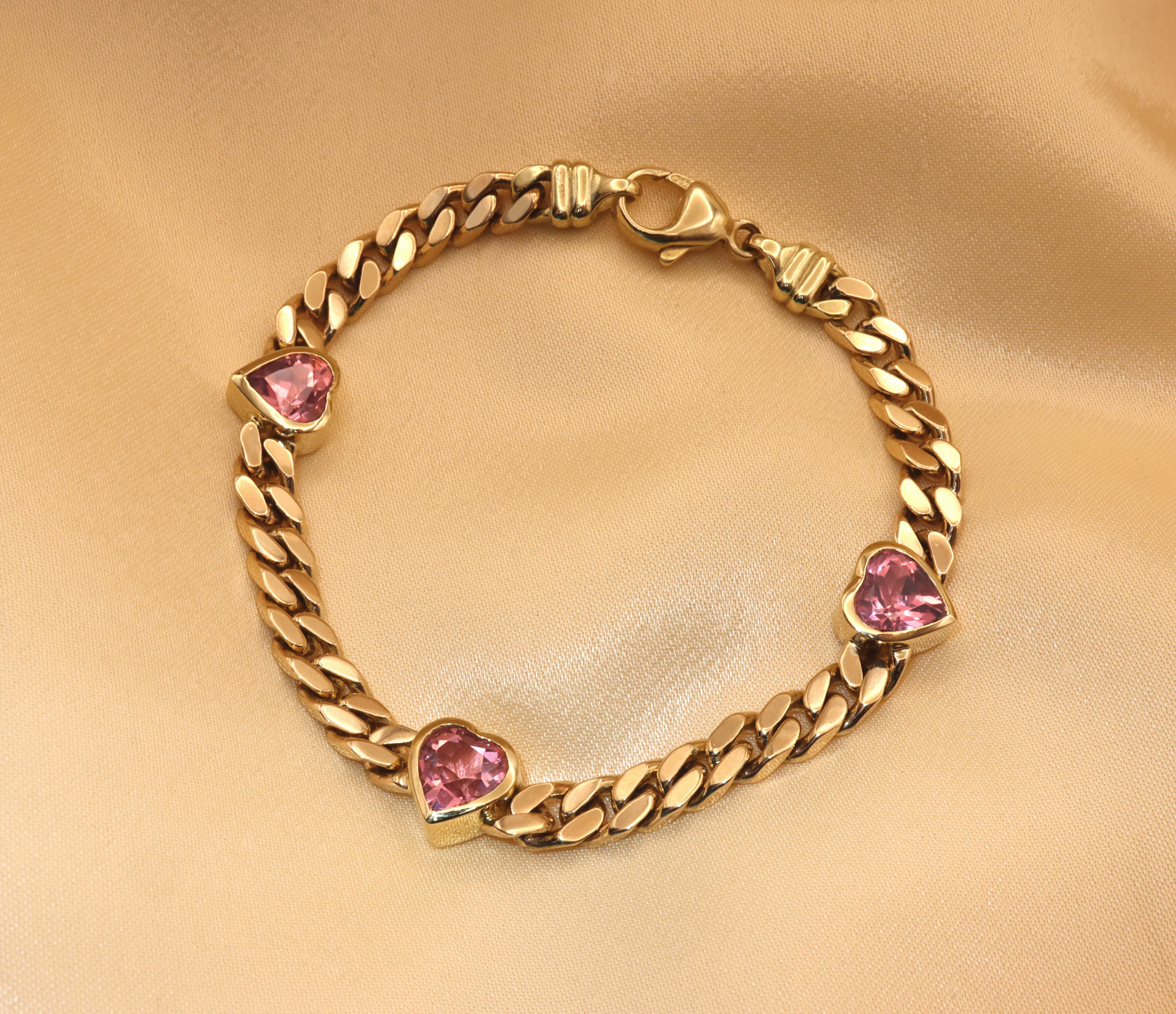 Goldene Panzerarmband mit rosa Turmalinherzen, detailreich, auf glänzendem Stoff liegend.
