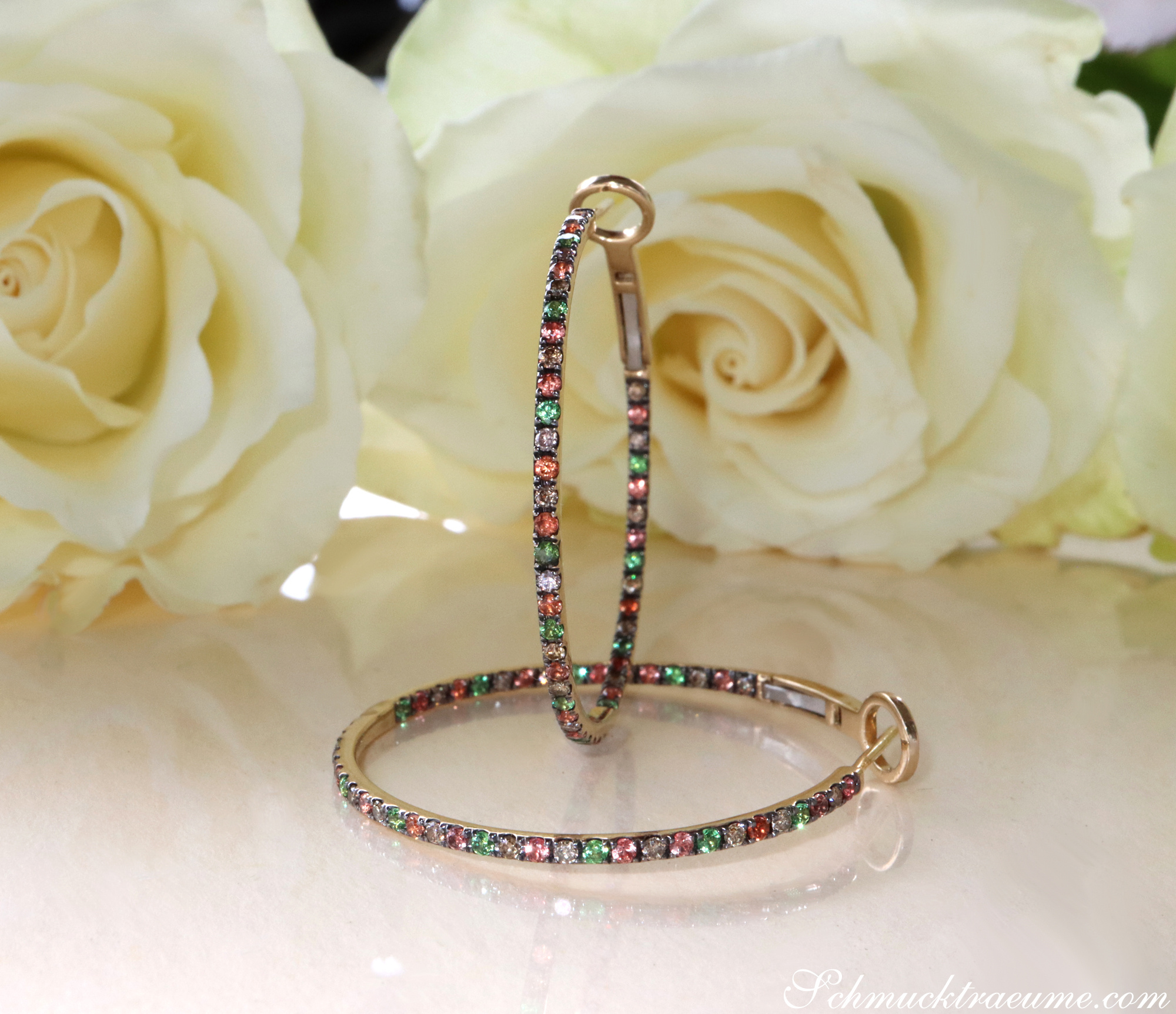 Goldene Creolen mit Tsavorite, braunen Diamanten und orangen Saphiren, Rosen im Hintergrund.