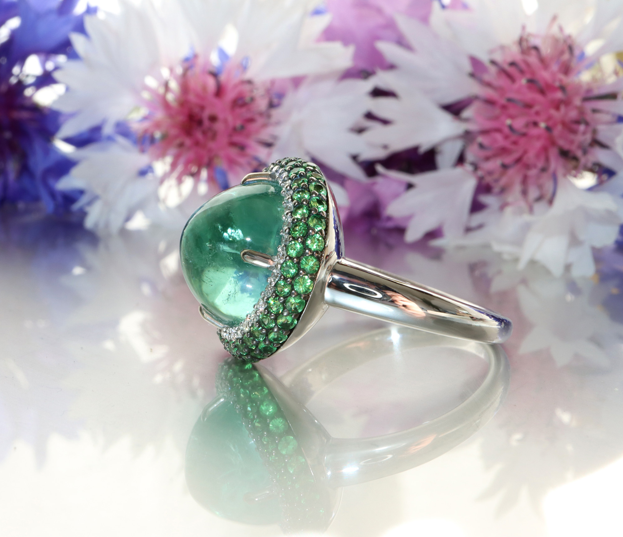 Mintfarbener Turmalin-Ring mit Tsavorite und Brillanten, Detailaufnahme, blühende Blumen im Hintergrund.