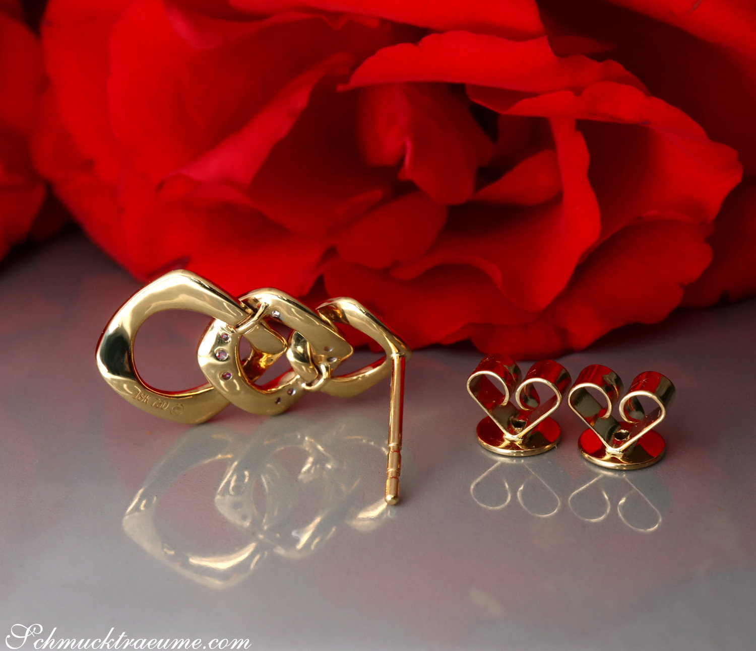 Goldene Ohrringe im Gliederdesign mit Brillanten, vor rotem Rosenblütenhintergrund.