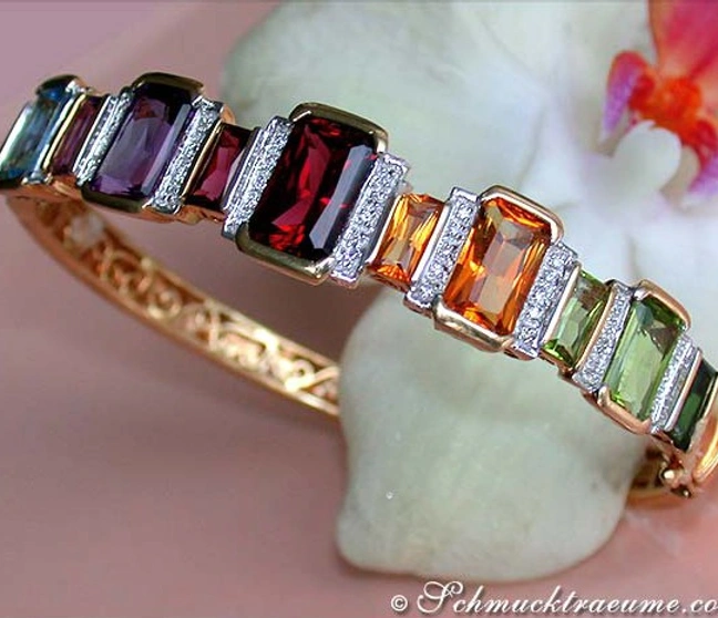 Multicolor Edelsteine und Diamanten auf Gelbgold-Armreif, Detailaufnahme mit Orchideenblüte im Hintergrund Multicolor Edelsteine und Diamanten auf Gelbgold-Armreif, Detailaufnahme mit Orchideenblüte im Hintergrund