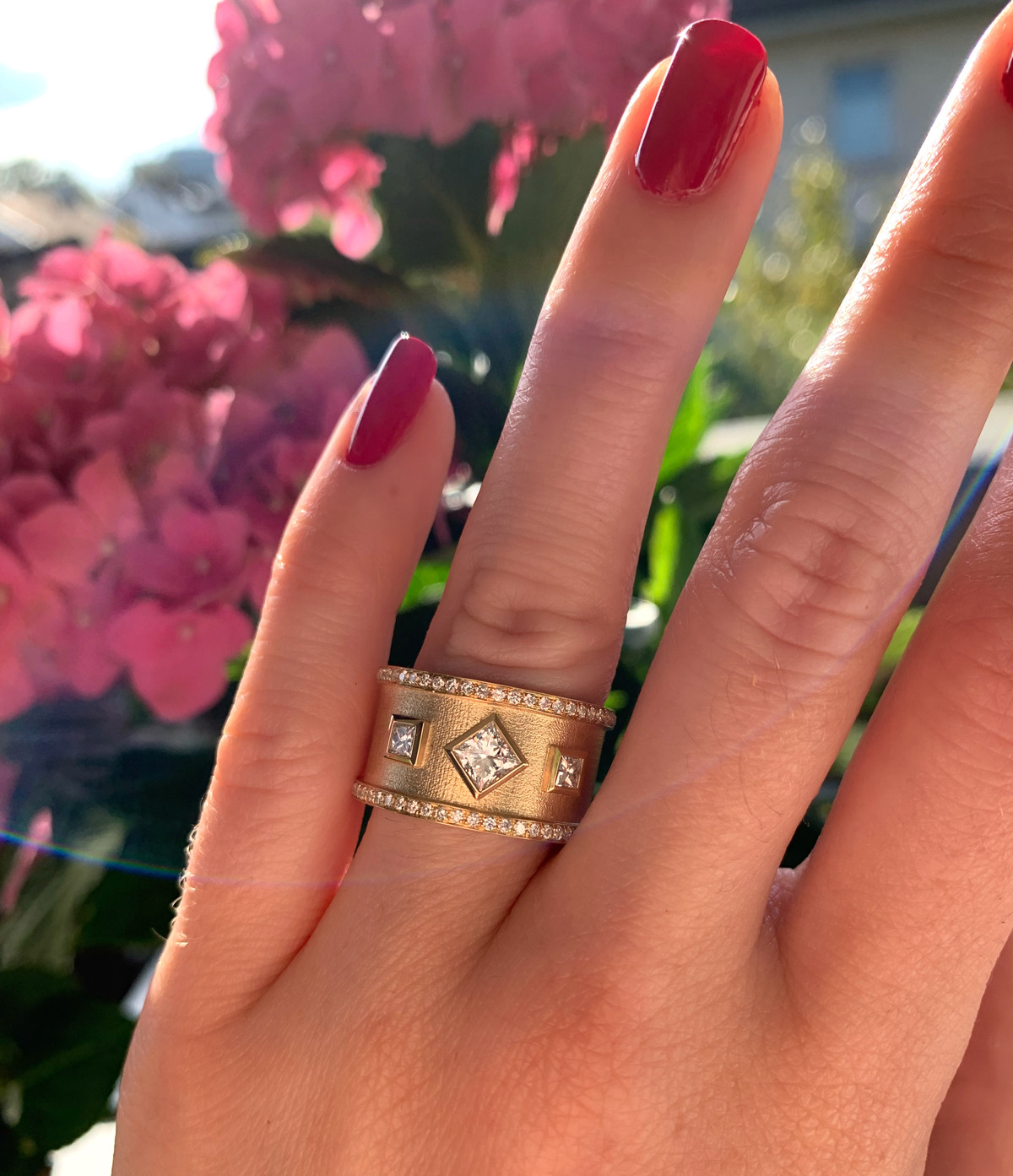Goldener Diamant-Ring mit Prinzessinen-Diamanten, Nagellack rot, Blumen im Hintergrund.