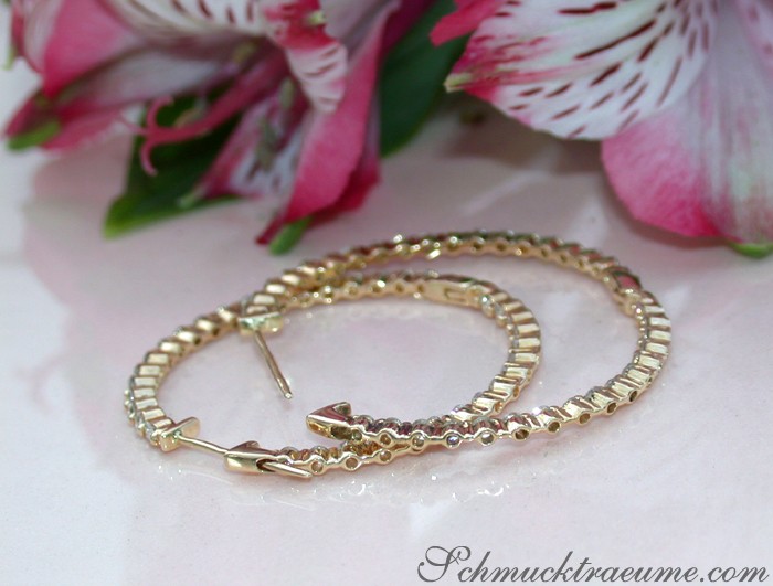 Goldene Creolen mit Brillanten auf weißem Untergrund, Blumen im Hintergrund.