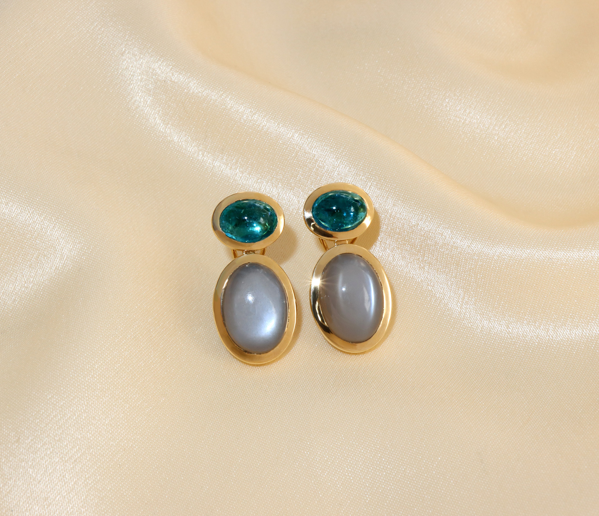 Goldene Ohrringe mit grünen und grauen Steinen auf cremefarbener Stoffoberfläche