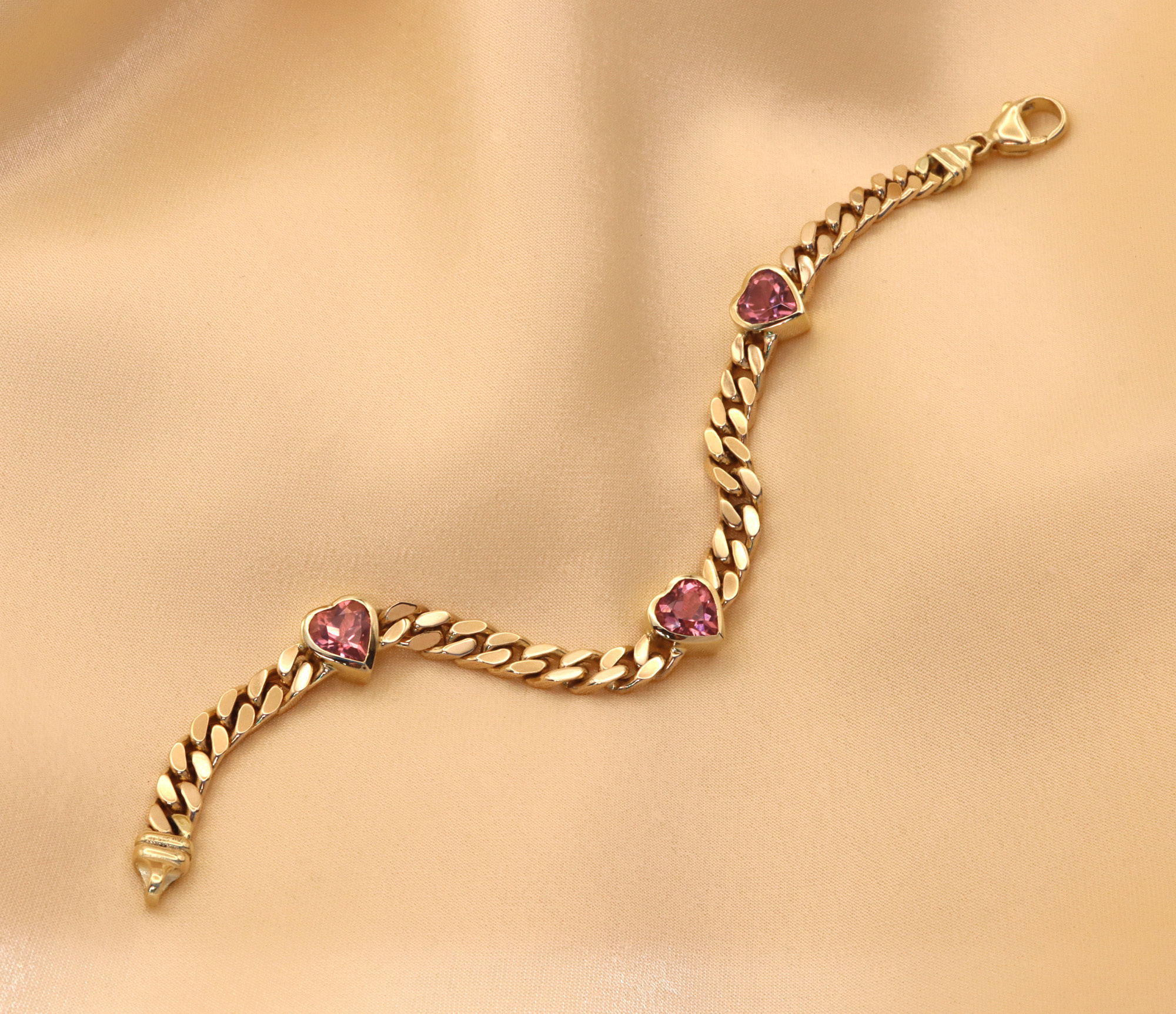 Goldene Panzerarmband-Detail mit rosa Turmalin-Herzen auf cremefarbener Seide.