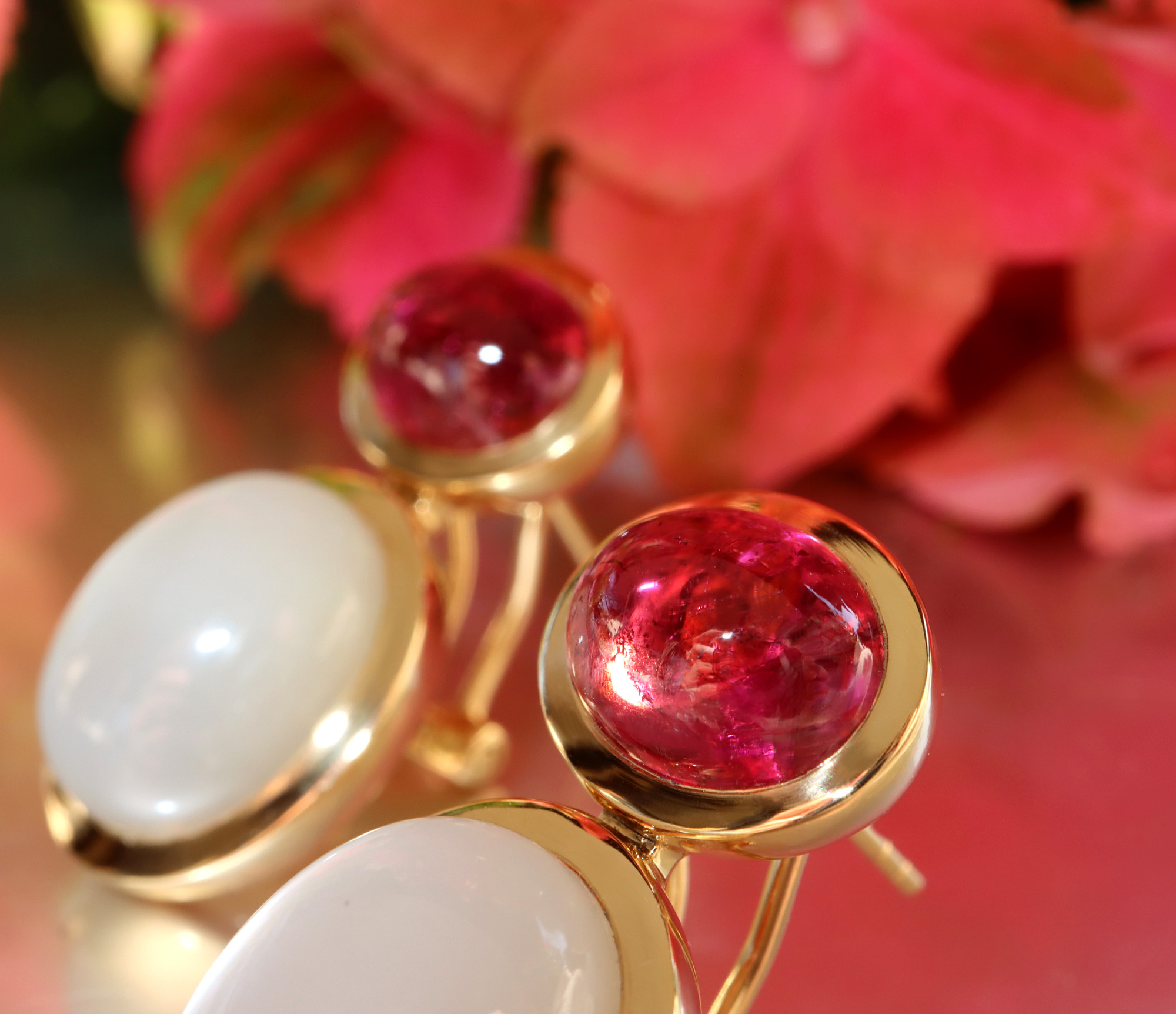 Goldene Ohrringe mit Rubellit und Mondstein, Detailaufnahme, sanfte Linien, elegant, vor Blütenhintergrund.