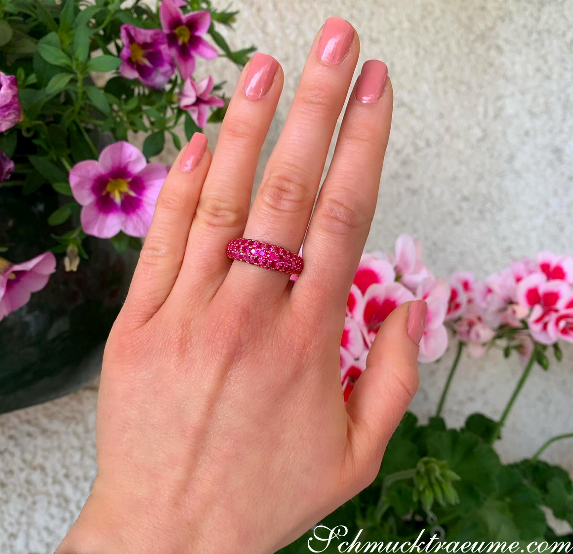 Rosenblüten im Hintergrund, handbesetzter Rubin Pavé Ring auf geöffnetem Finger, Nagellack in Rosa.