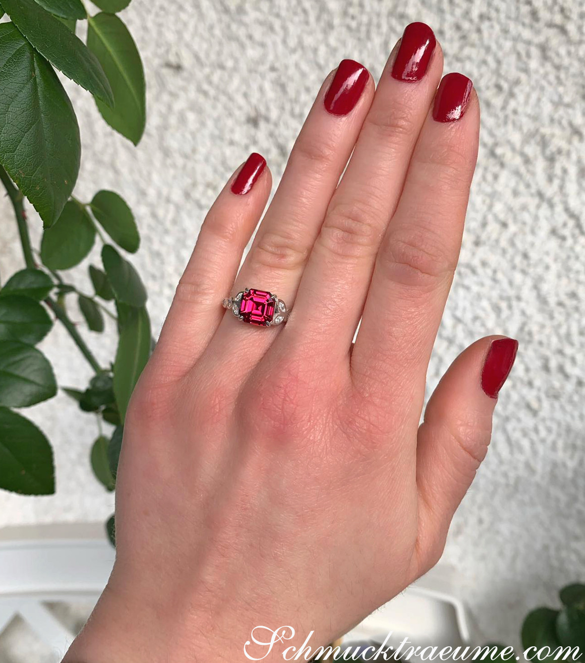 Sehr eleganter Rhodolith-Ring mit Diamanten auf der Hand, Nagellack rot, Pflanzen im Hintergrund.