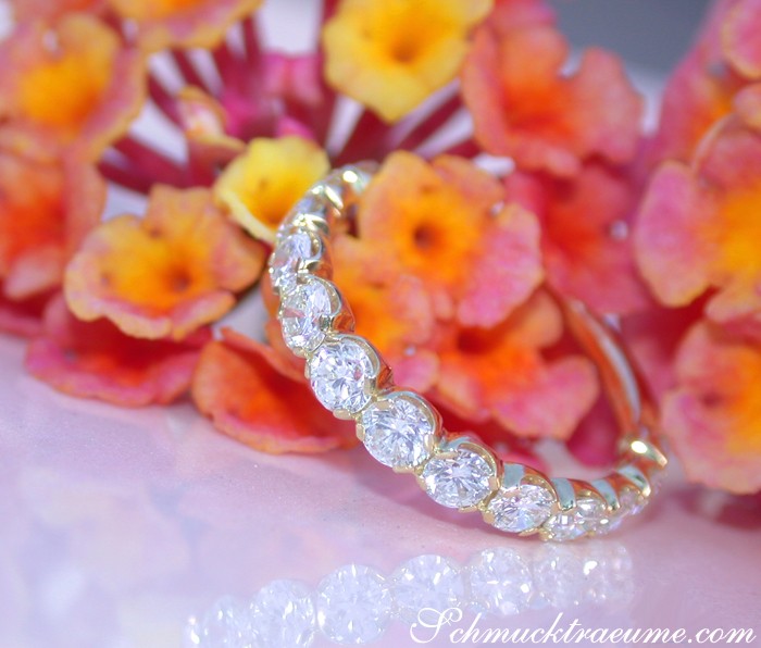 Goldener Memory Ring mit ovalen Brillanten, detailreich, vor farbenfrohen Blüten.