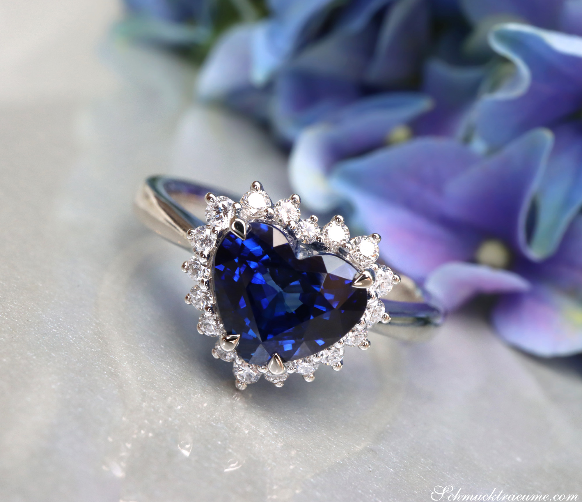 Herzform-Saphir-Ring mit Brillanten in Weißgold 750, Detailaufnahme, blauer Saphir, Diamantenbesatz, Blume im Hintergrund