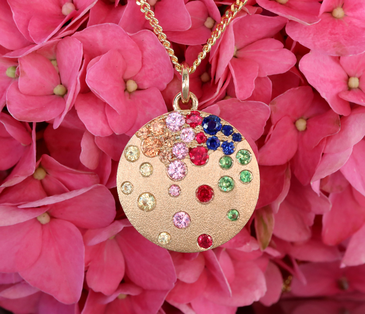 Goldene Medaillon-Halskette mit farbenfrohen Saphiren, Rubinen und Tsavorites auf rosa Hortensienblüten.