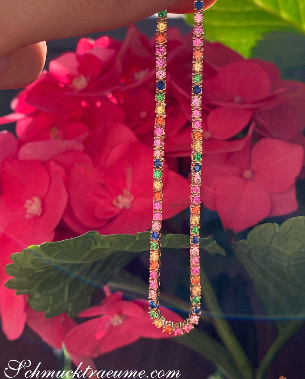 Multicolor-Saphir-Tennisarmband mit feinen Steinen in Gelbgold, vor roten Blüten.