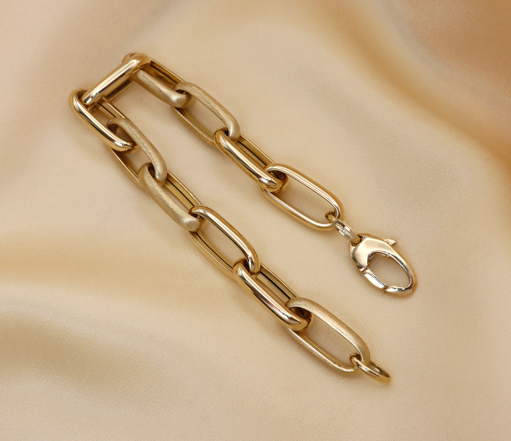 Goldene Paperclip-Armband mit glänzenden und satinierten Elementen auf cremefarbener Stoffoberfläche