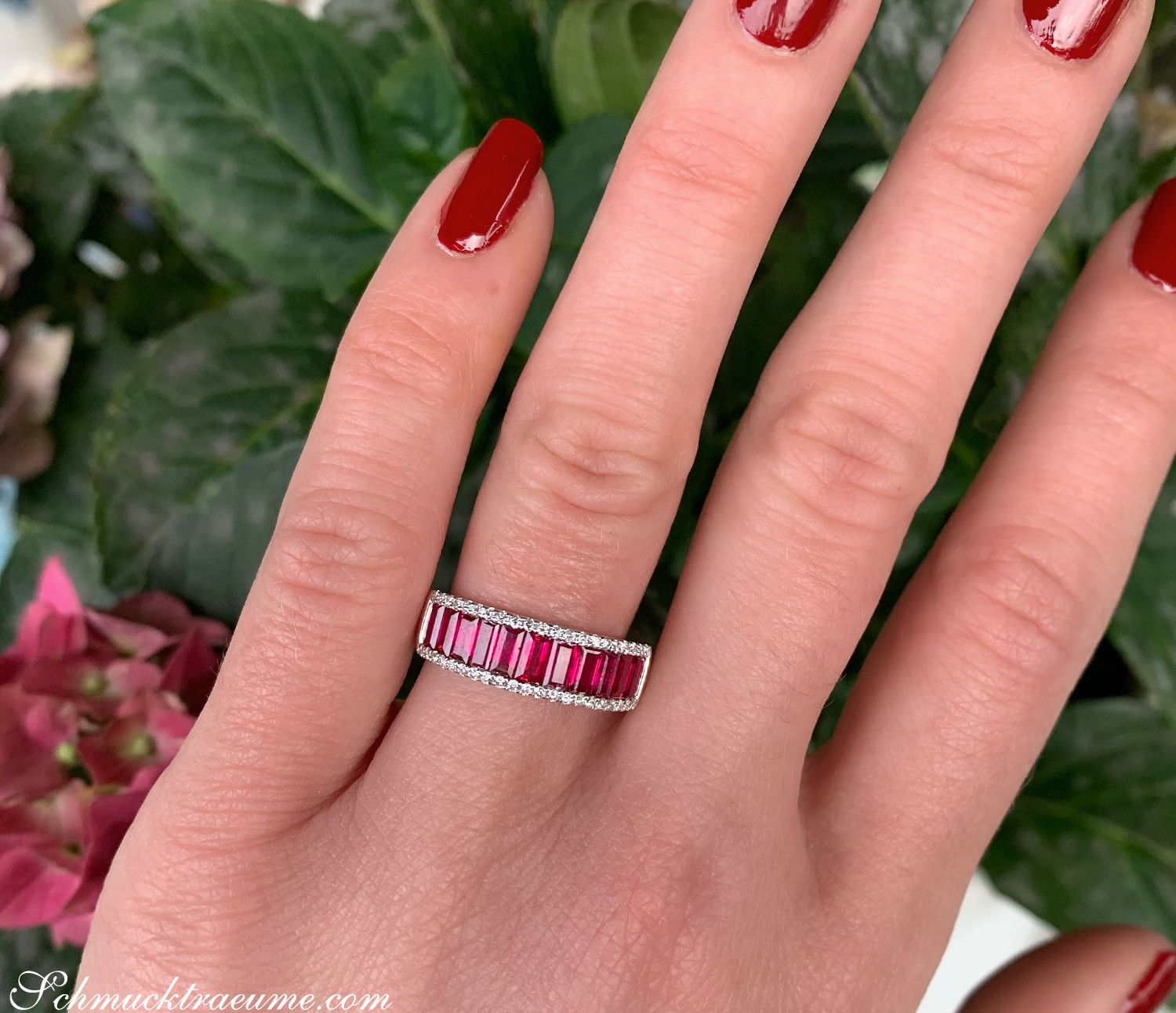 Rosenrot lackierte Nägel, weißgoldener Ring mit Baguette-Rubinen und Diamanten, Hintergrund mit grünen Blättern.