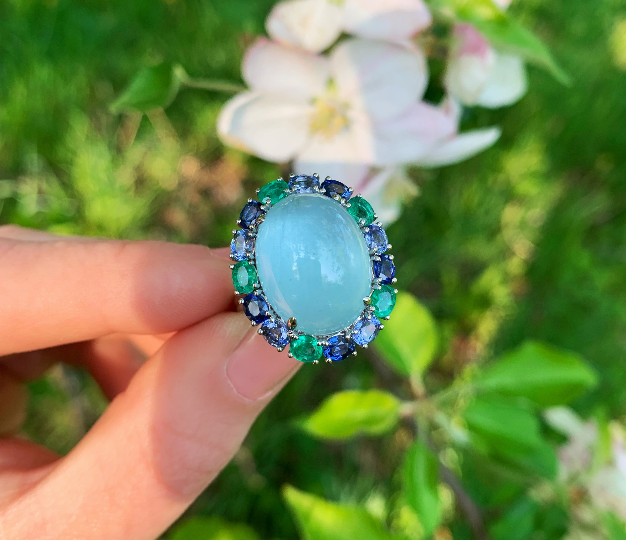 Milky Aquamarin-Ring mit Saphiren und Smaragden im Cabochon-Stil, Hintergrund mit Blüten.