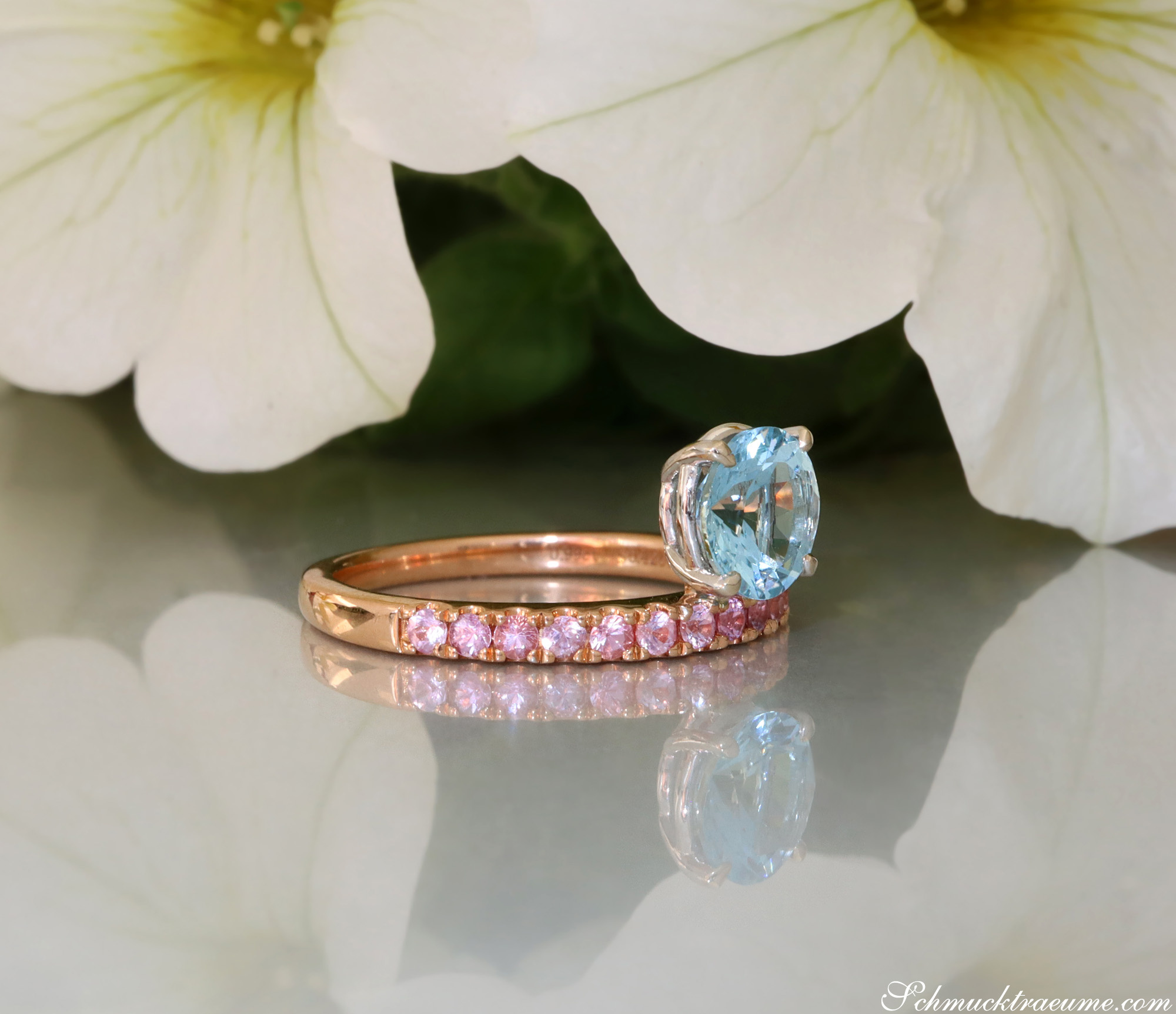 Weißgold-Ring mit Aquamarin und rosa Saphiren, Detailaufnahme auf glänzender Oberfläche, Blüte im Hintergrund.