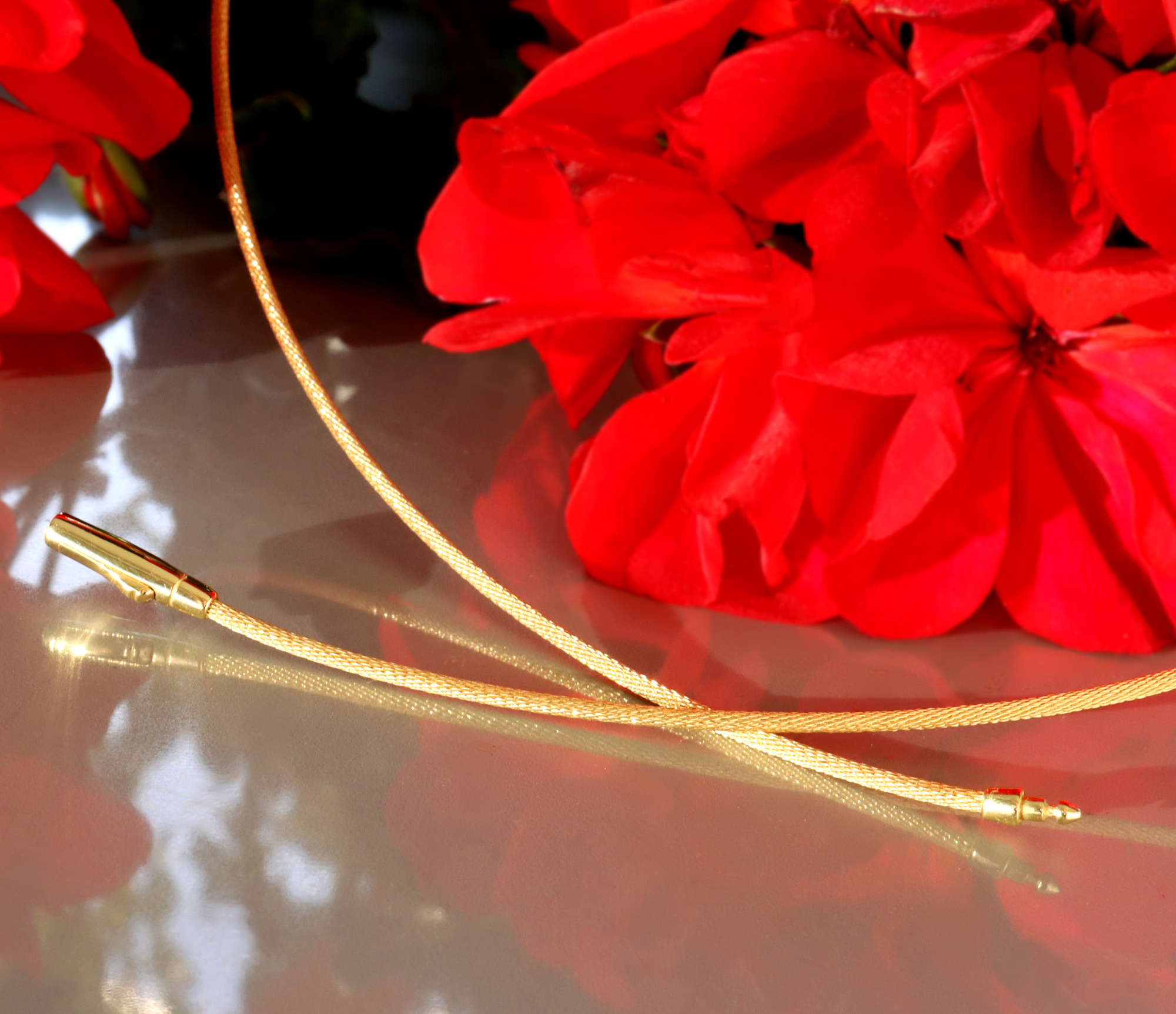 Gold Pur: Flüssig geflochtener Halsreif in Gelbgold 750, Detailaufnahme mit roten Blüten im Hintergrund