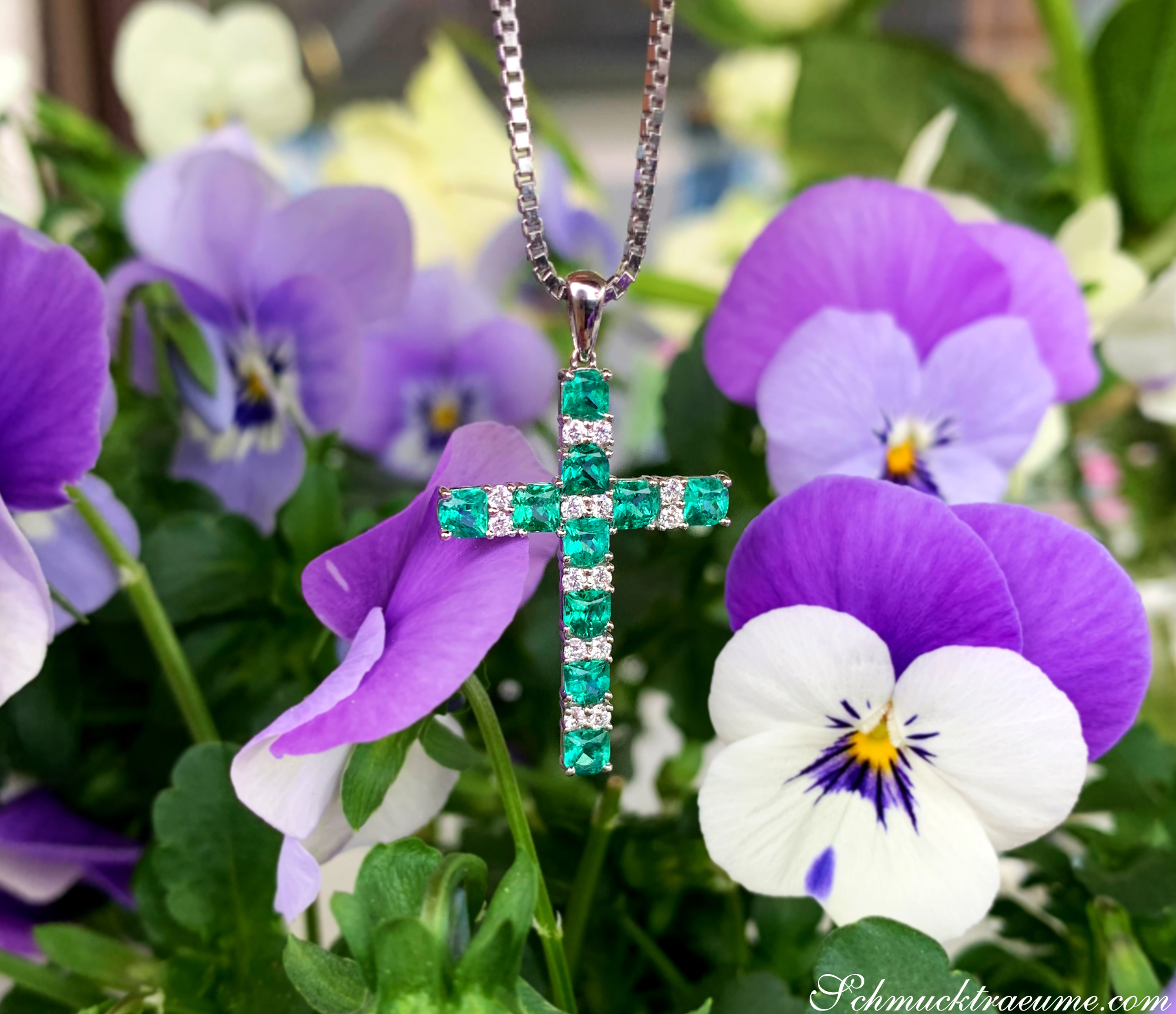 Blütenpracht: Smaragd-Kreuz-Anhänger mit Brillanten, umgeben von lila und weißen Blüten.