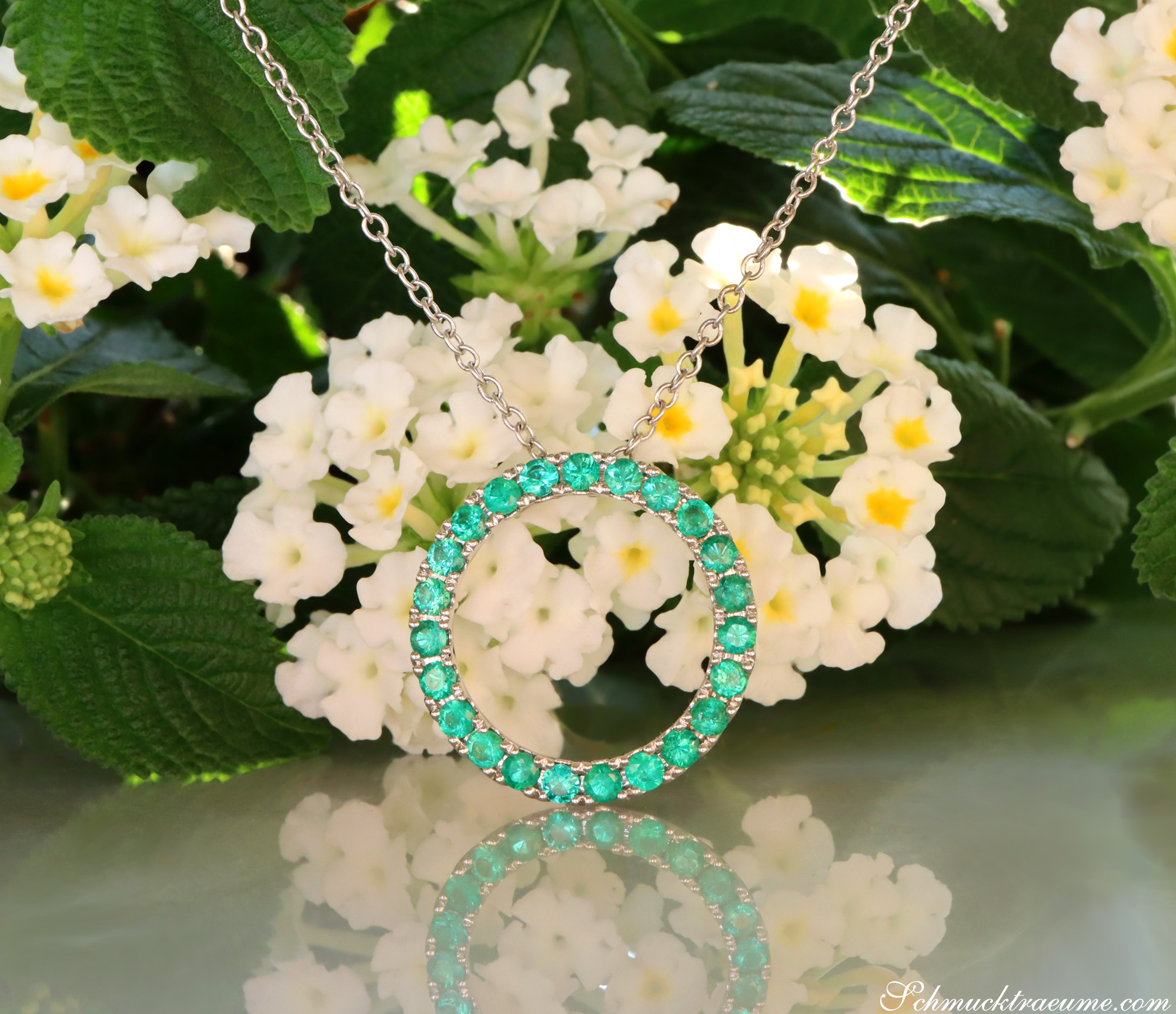 Smaragd Kreisanhänger in Weißgold, umgeben von weißen Blumen und grünen Blättern.
