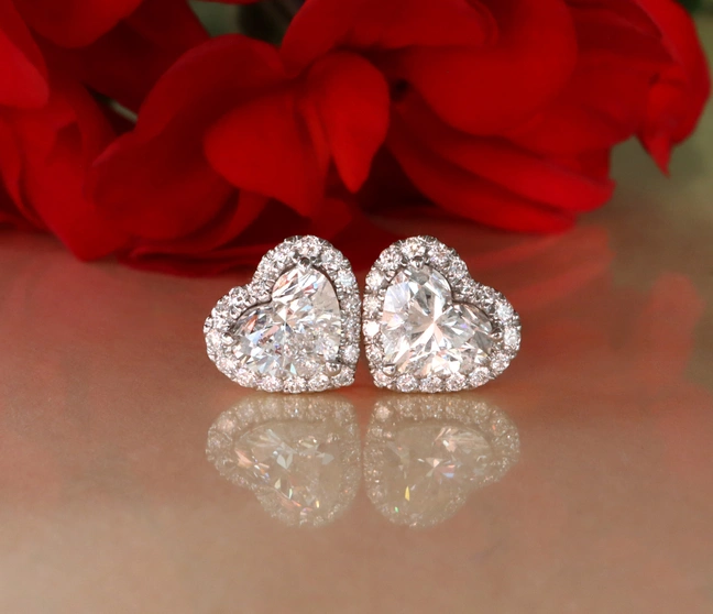 Herzformige Diamant-Ohrstecker mit Brillanten, 1,20 ct., Weißgold 750, vor rotem Rosenblütenhintergrund. Herzformige Diamant-Ohrstecker mit Brillanten, 1,20 ct., Weißgold 750, vor rotem Rosenblütenhintergrund.